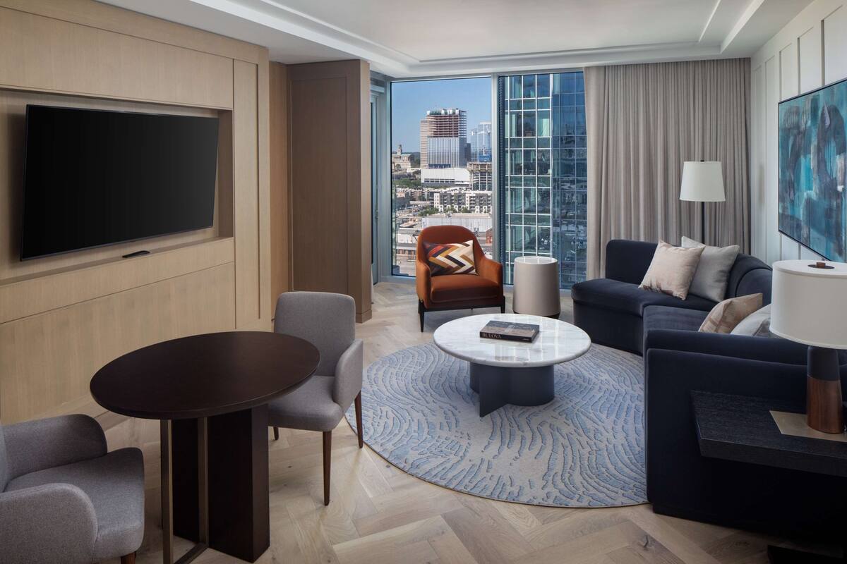 A modern hotel room with a large flat-screen TV, a round coffee table, a sofa, and a chair. The room features a large window with a city view.