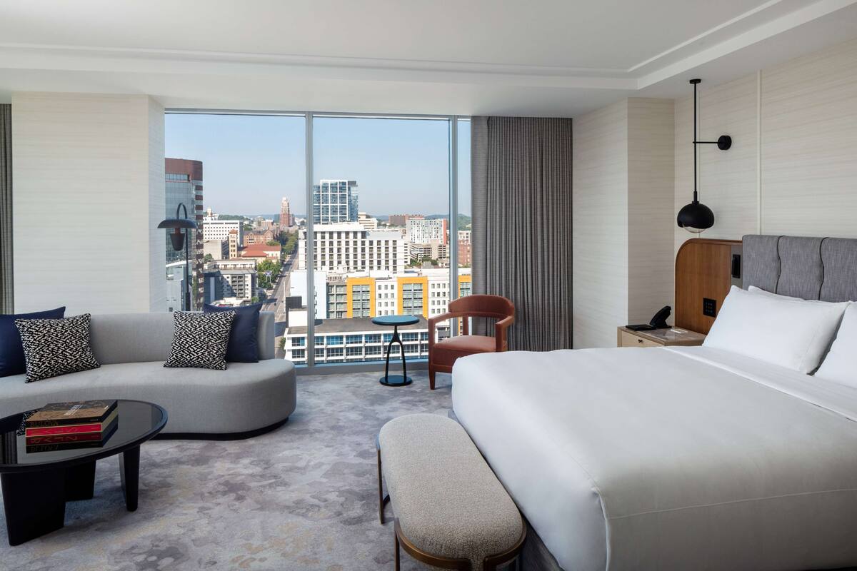 A modern hotel room with a large bed, a sofa, a small table, and a chair. The room has a large window with a city view.