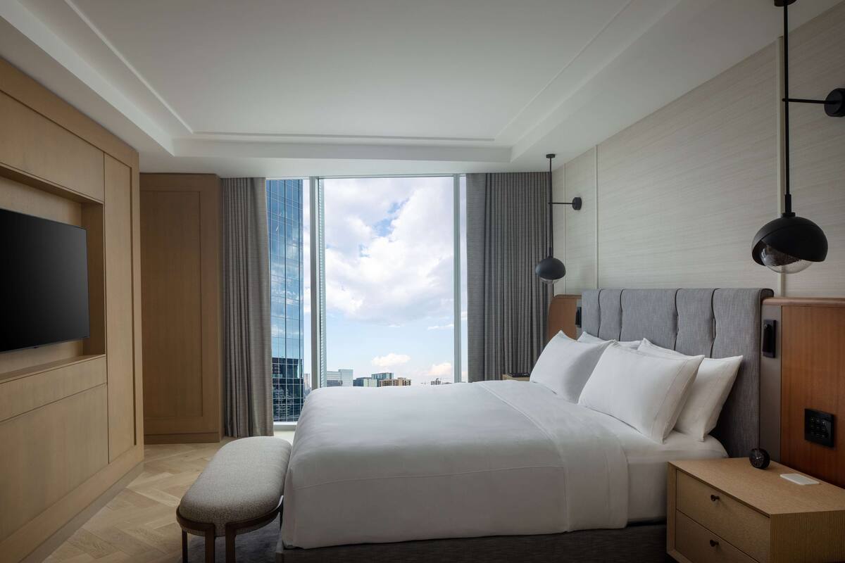 A modern hotel room with a large bed, a flat-screen TV, a bedside table, and a view of the city skyline.