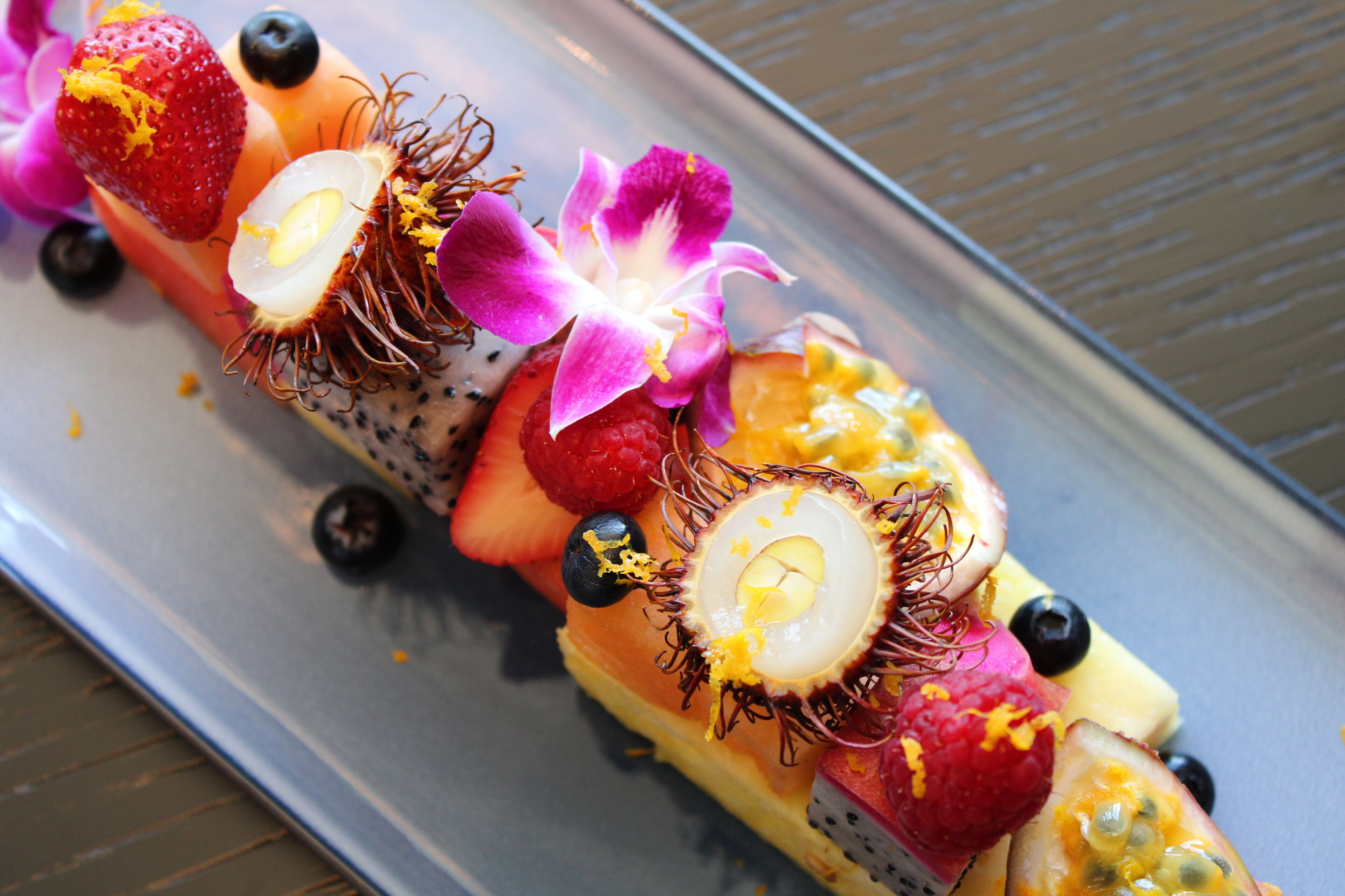 Un postre con diversas frutas y una flor sobre una bandeja.