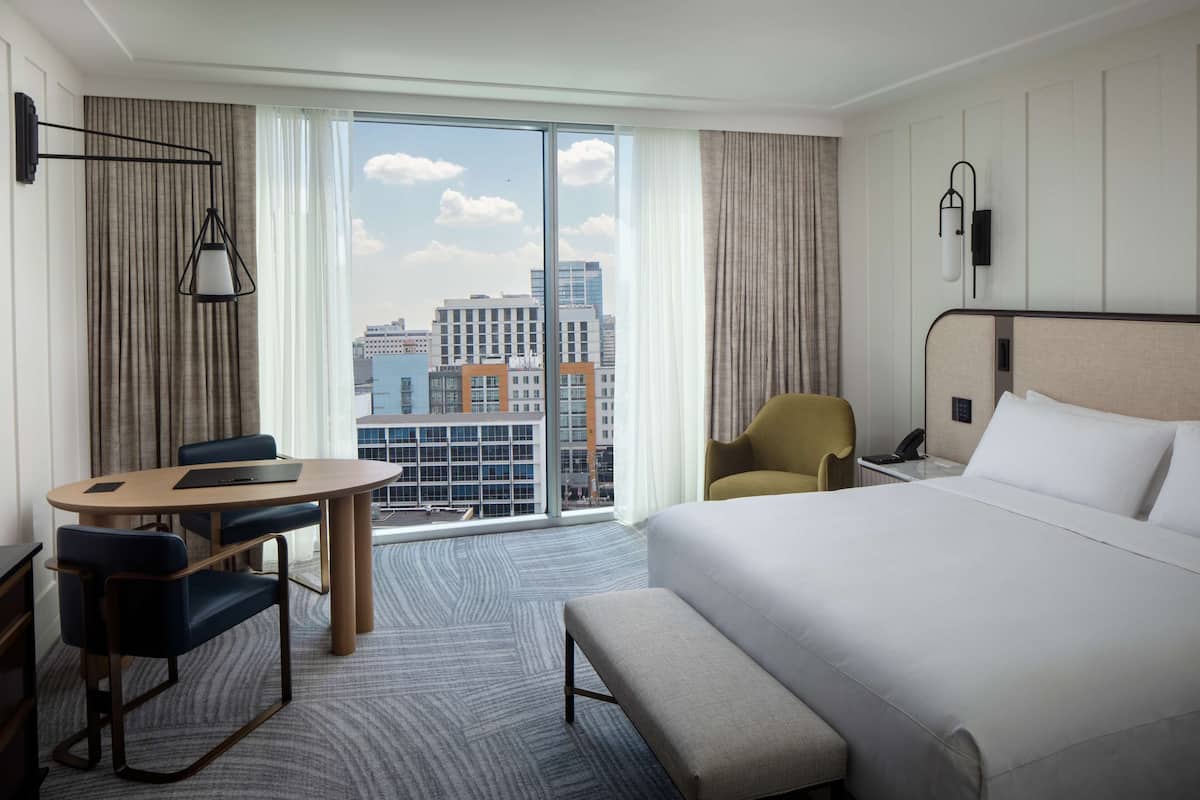 A hotel room with a large bed, a desk with chairs, a view of the city through floor-to-ceiling windows, and a cityscape visible outside.