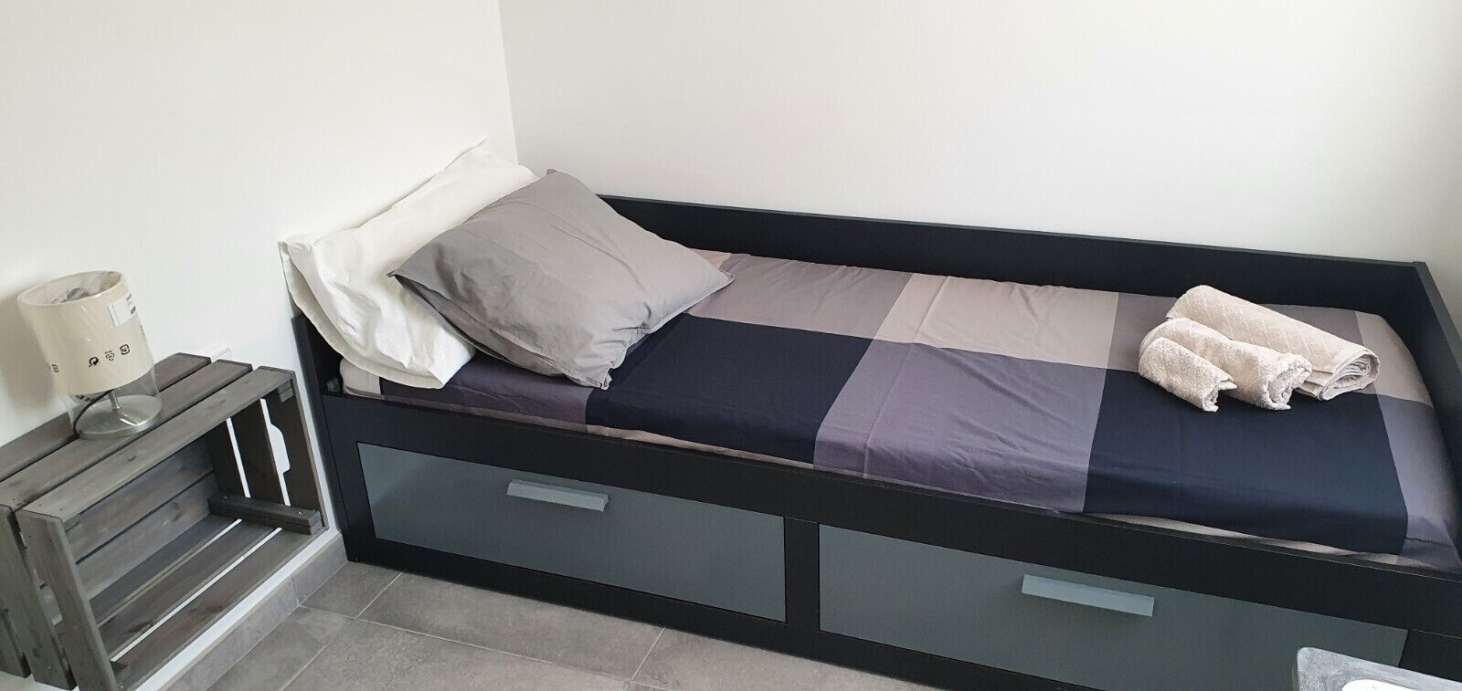 A single bed with a dark frame, grey and blue bedding, and a folded towel on the right side.