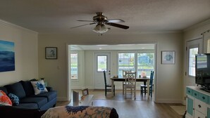 A living room with a dark blue sofa, a ceiling fan, a dining area with a table and chairs, and a kitchen area with cabinets. - Gulf of Mexico Canal Cottage with Boat Dock and Heated Pool (New Port Richey)