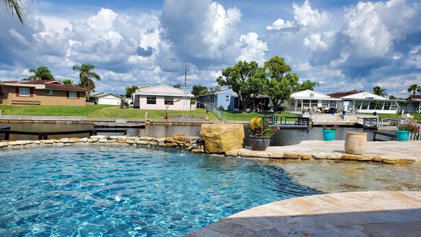 A swimming pool with clear water, surrounded by a stone deck. In the background, there are houses with red roofs and a grassy area. - Gulf of Mexico Canal Cottage with Boat Dock and Heated Pool (New Port Richey)