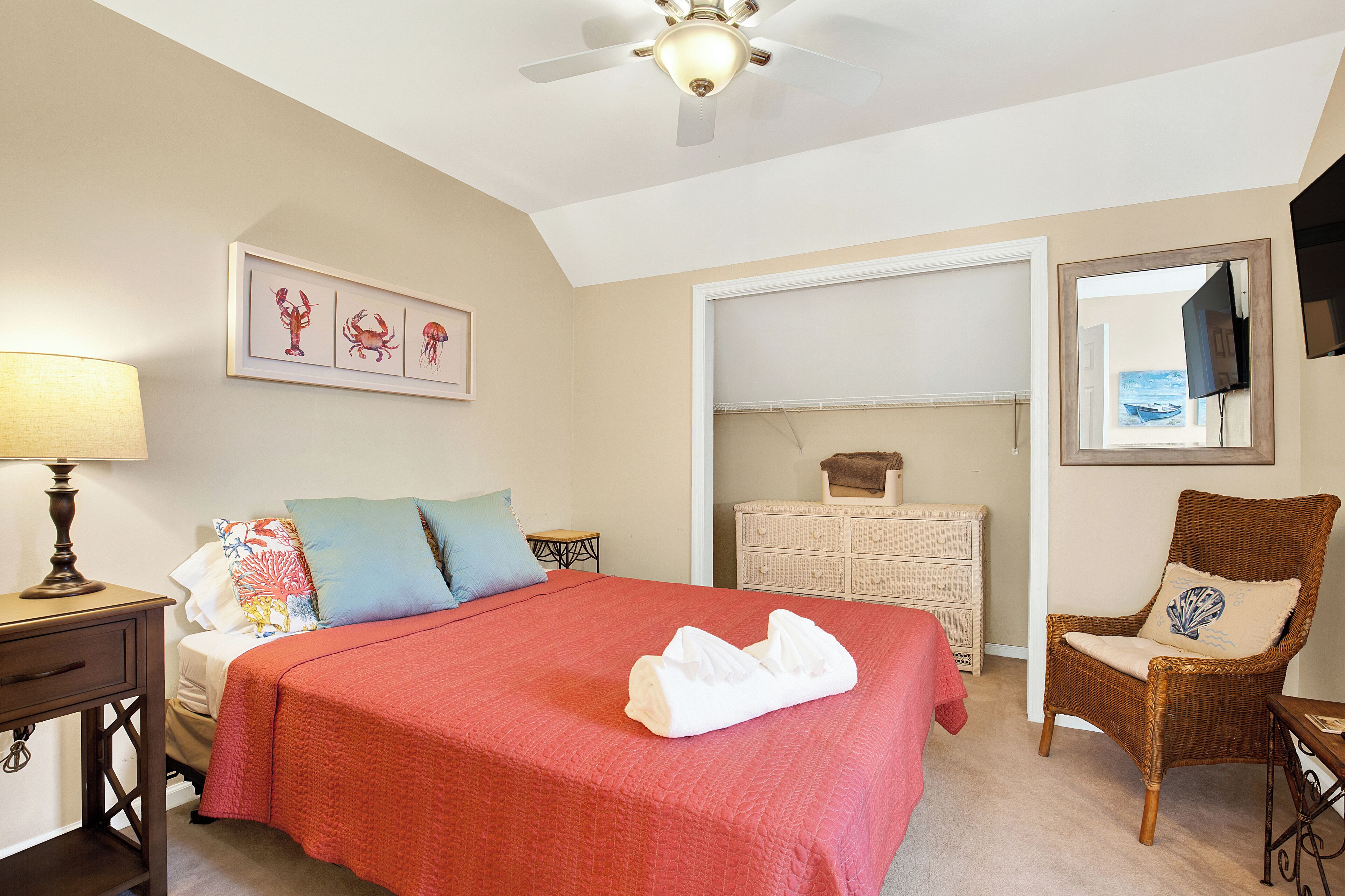 Une chambre à coucher comprenant un lit, une table de chevet, une commode, une chaise, un ventilateur de plafond et un tableau encadré accroché au mur.