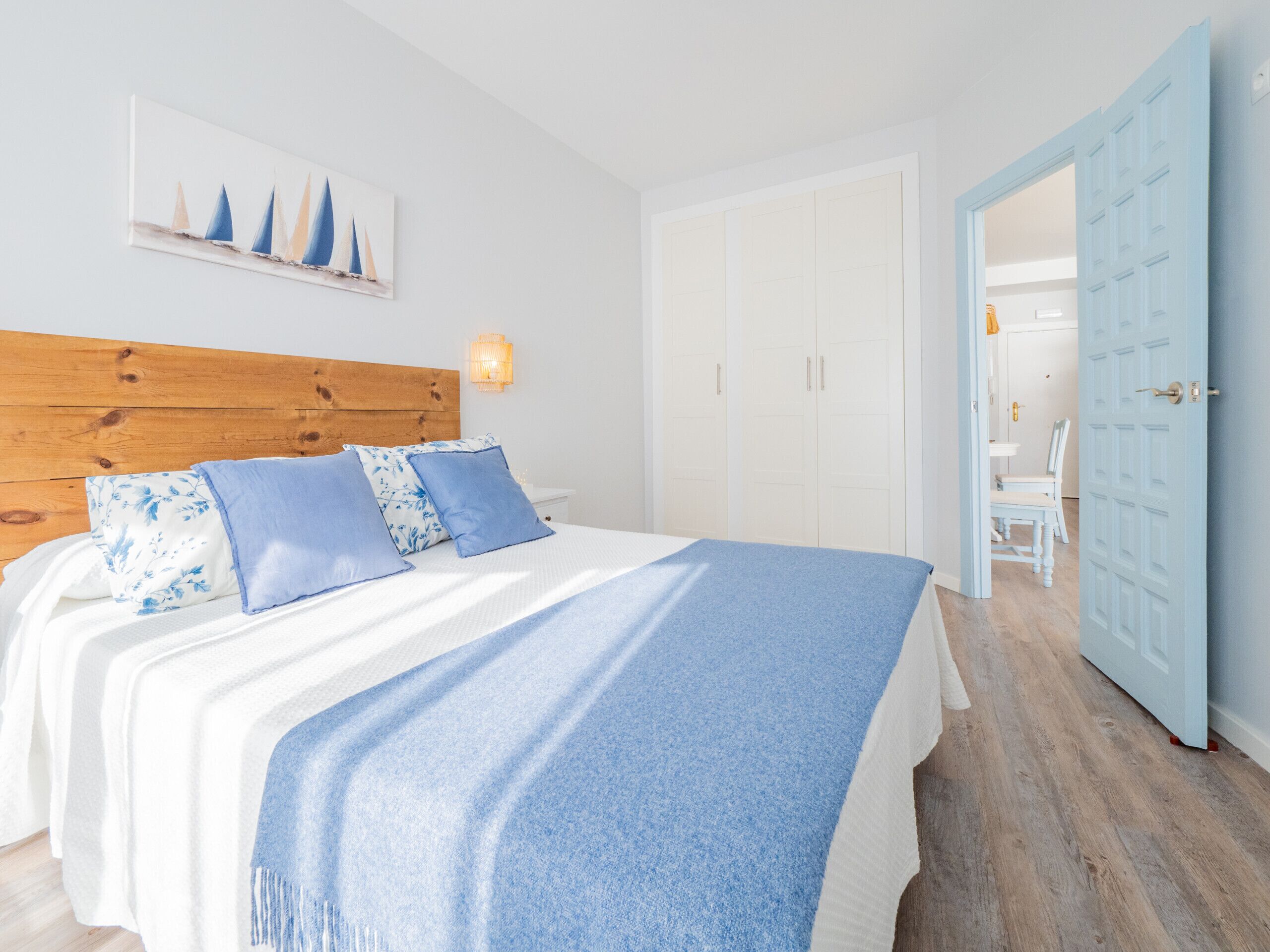 Une chambre à coucher avec une tête de lit en bois, un lit recouvert d’une courtepointe bleue et blanche, et un tableau représentant des voiliers accroché au mur.