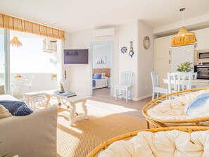 A bright, airy living room with a sofa, a dining table, and a kitchen area with a TV and clock. - Cubo’s La Bambera First Line Beach / sea views / free wifi (Mijas Costa)