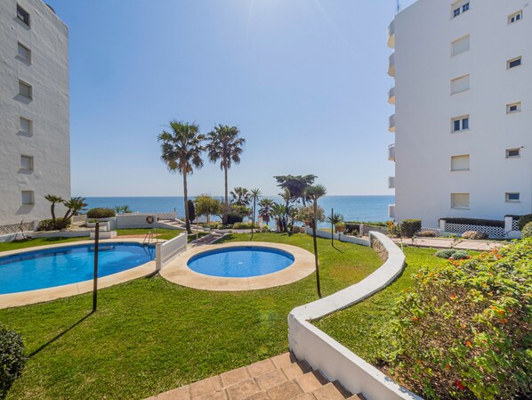 Uma área de piscina com duas piscinas, cercada por vegetação e palmeiras, com vista para o mar.