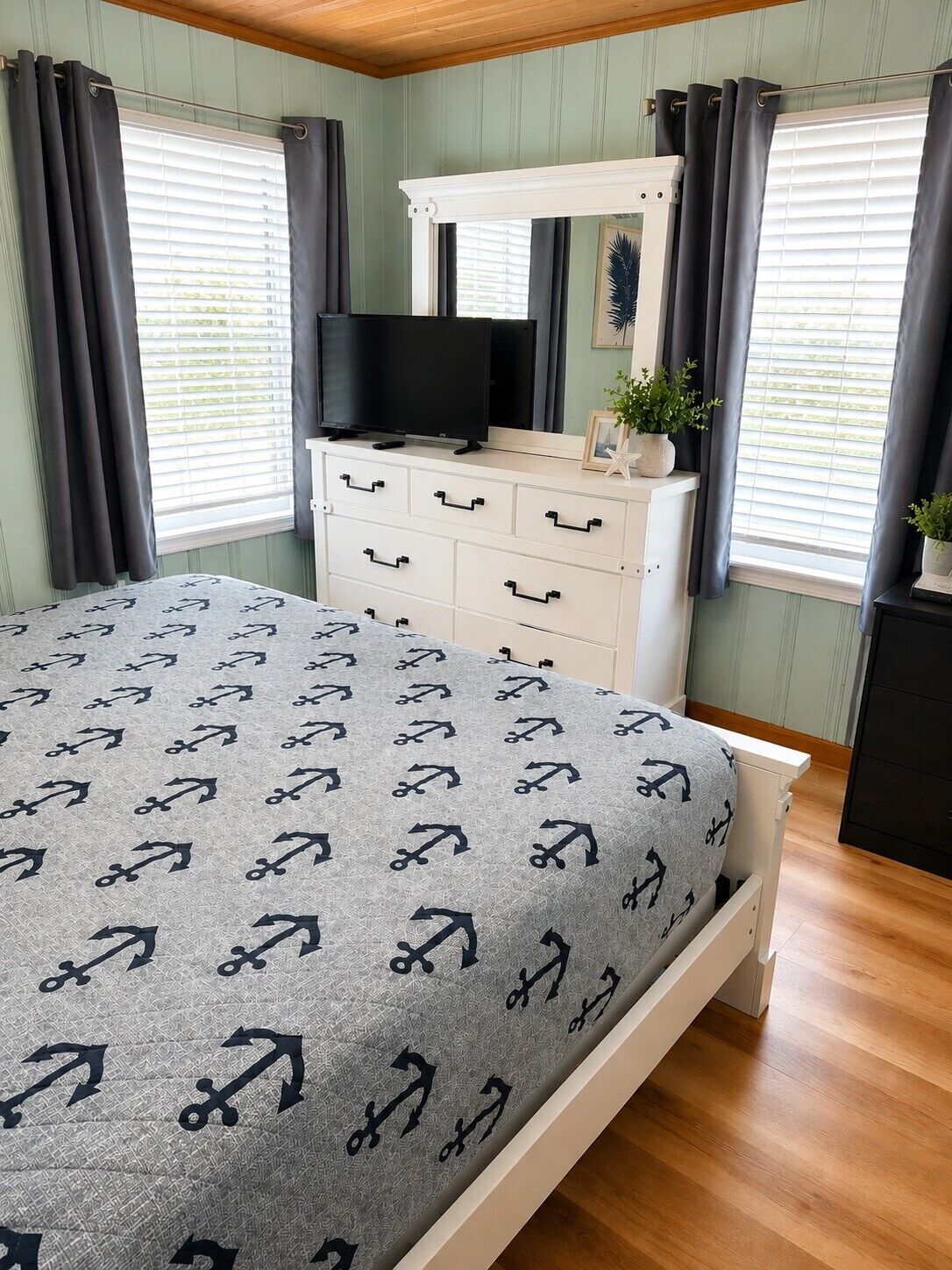 A bedroom with a bed, a TV on a dresser, and two windows with curtains.