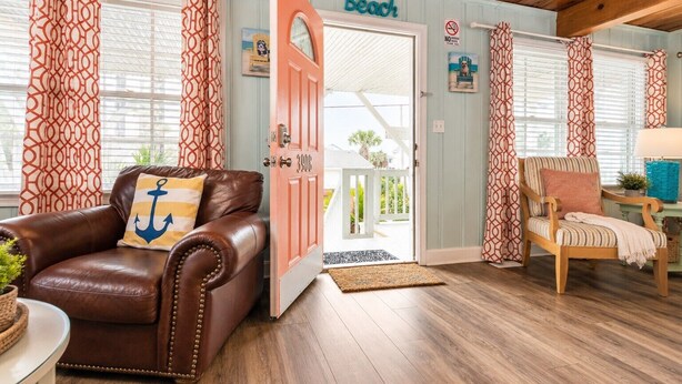Una habitación con un sofá de cuero marrón, una silla de madera, una puerta con la etiqueta 'Beach' y vistas al exterior.