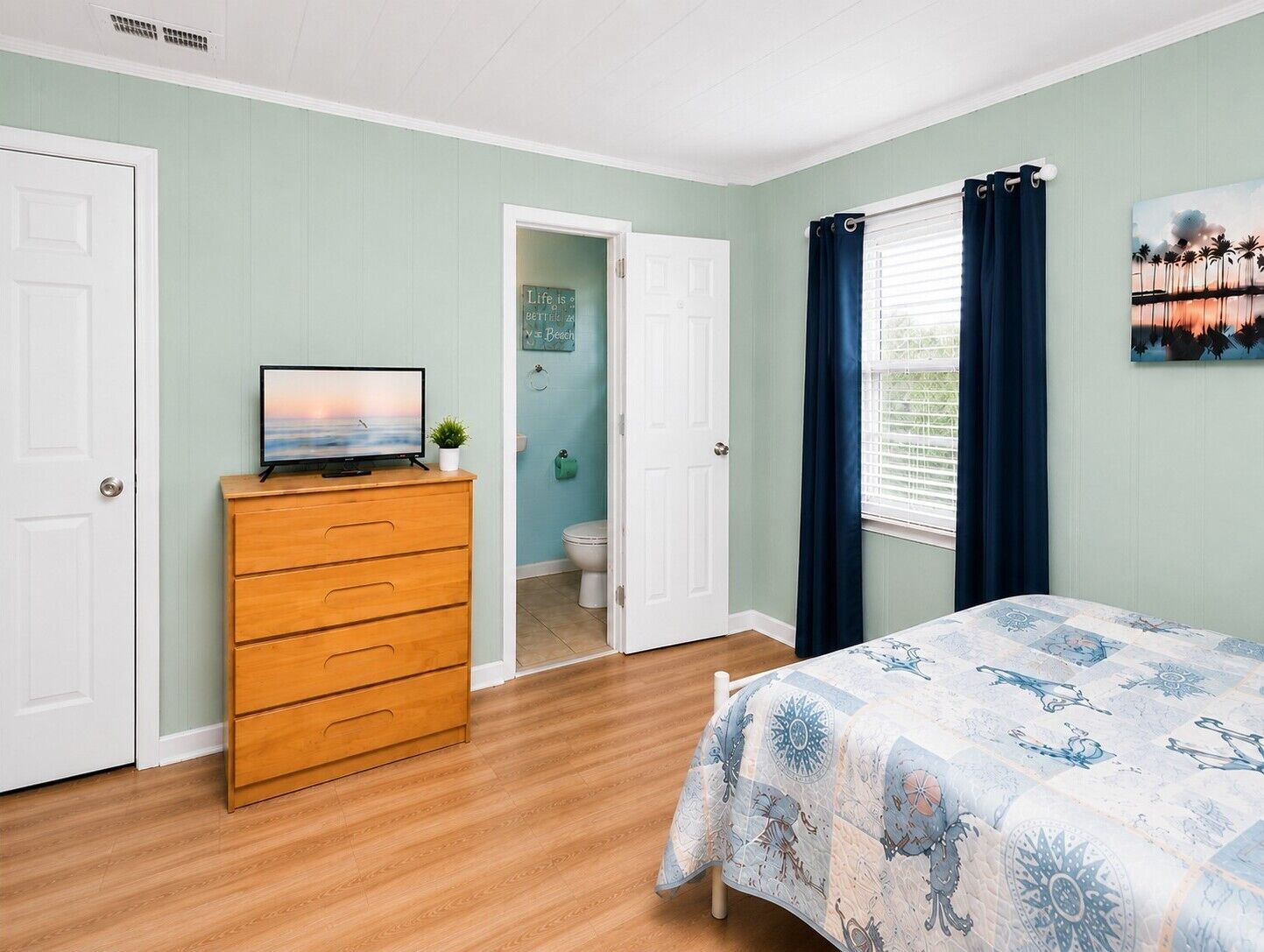 Un dormitorio con cama, cómoda, televisor sobre la cómoda, ventana con cortinas y puerta que da a un baño.