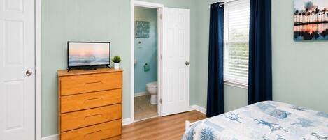 Un dormitorio con cama, cómoda, televisor sobre la cómoda, ventana con cortinas y puerta que da a un baño.