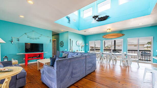 A spacious living room with a blue sofa, wooden flooring, and a TV on a red stand. - Bring your Big Group, 12 BR/8 BA, Private Pool, Walk to the Beach! (Port Aransas)