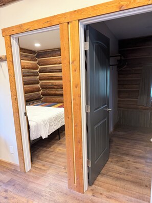 A room with a bed, wooden walls, and a door leading to another area. - Teton Cabin at Columbia Mountain Ranch (Columbia Falls)