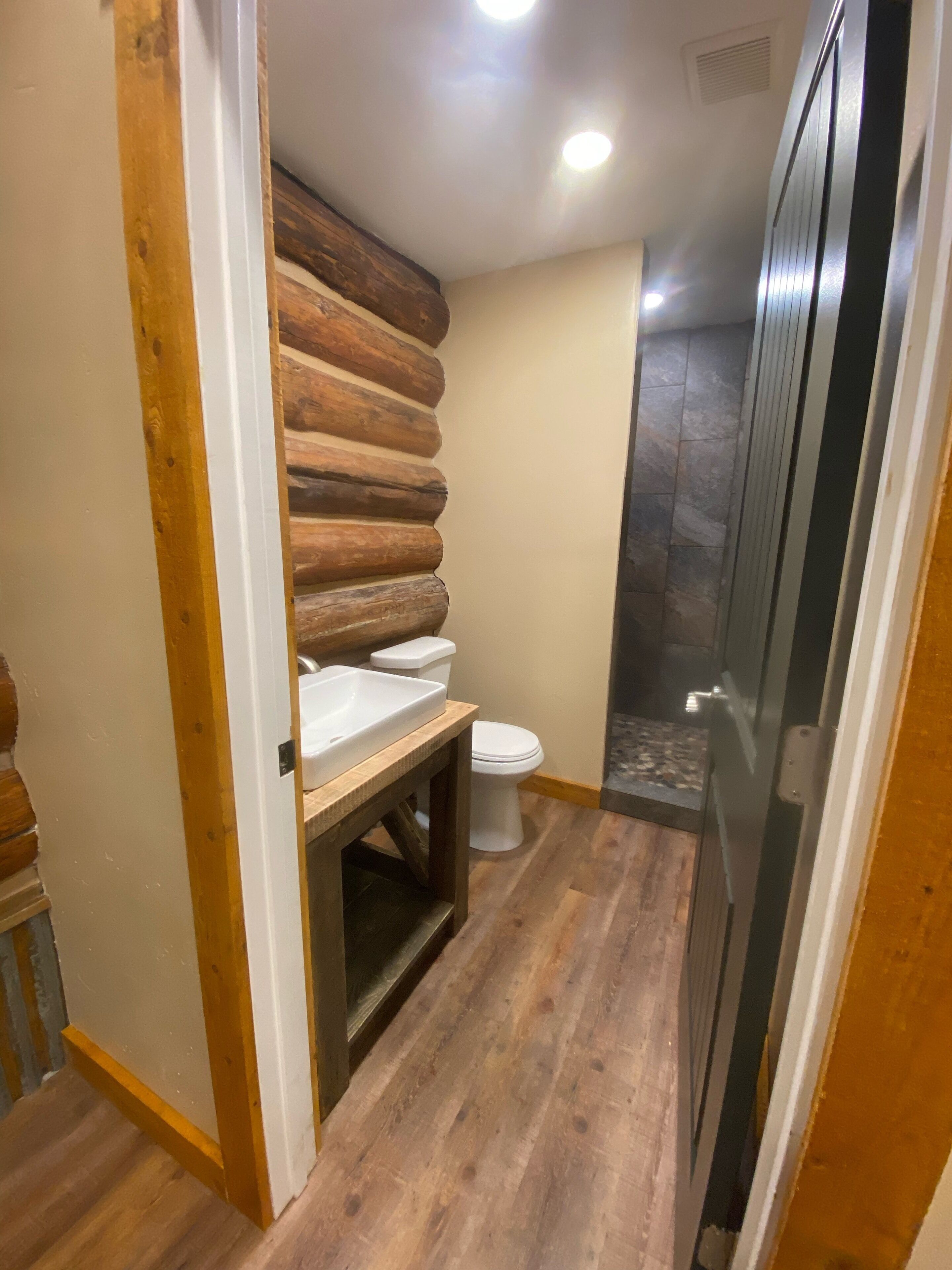 A bathroom with a wooden log wall, a white sink, a toilet, and a shower area.