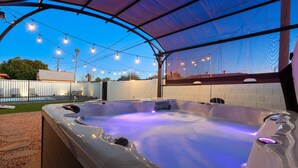 Un jacuzzi bajo una estructura cubierta, con luces colgantes, rodeado de un muro de ladrillo y una piscina al fondo.