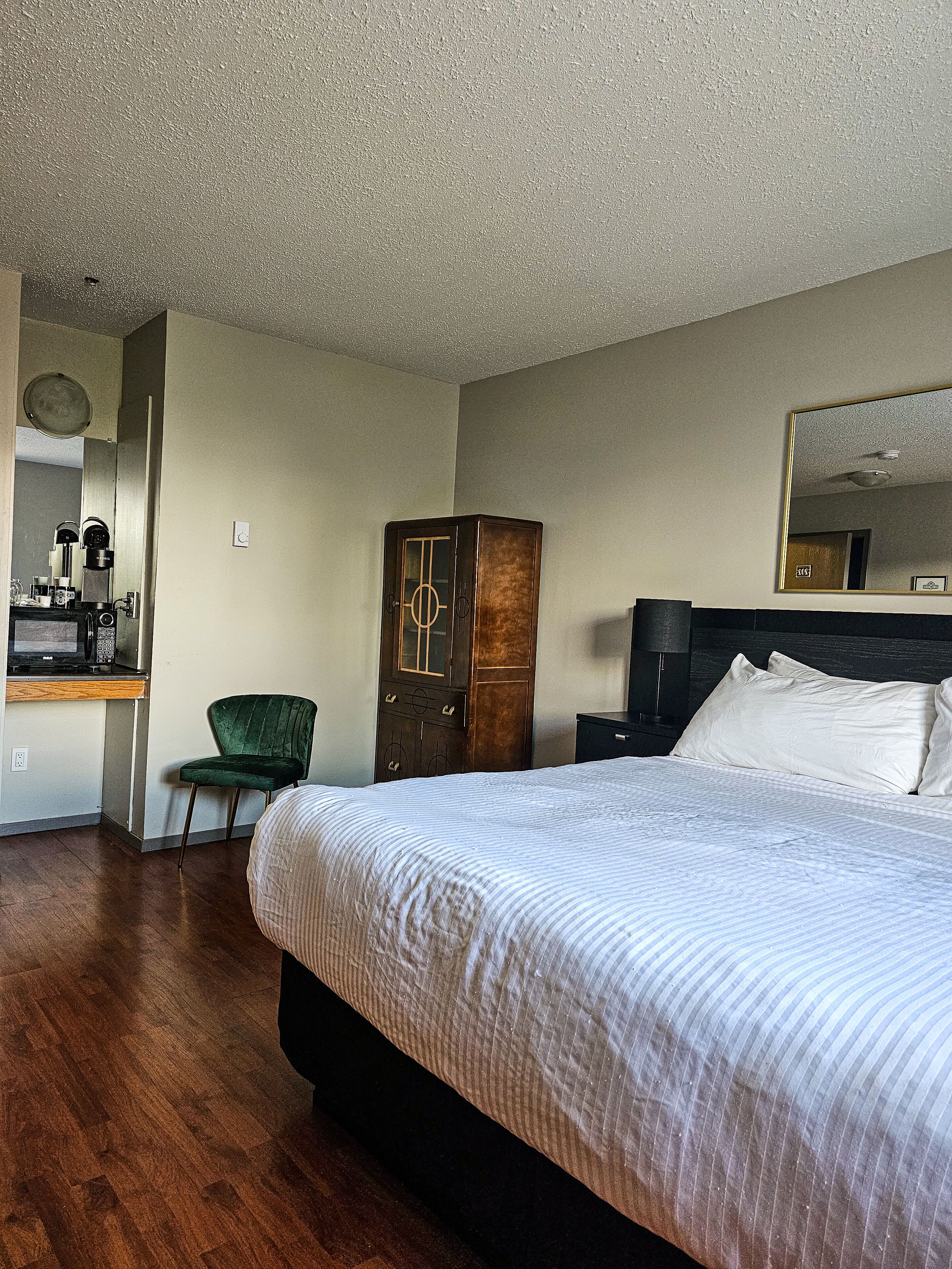A hotel room with a large bed, a wooden wardrobe, a green chair, a desk with a coffee maker, and a mirror on the wall.