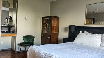 A hotel room with a large bed, a wooden wardrobe, a green chair, a desk with a coffee maker, and a mirror on the wall.