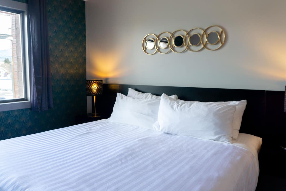 A neatly made bed with white linens, a wall-mounted circular mirror, and a bedside lamp.