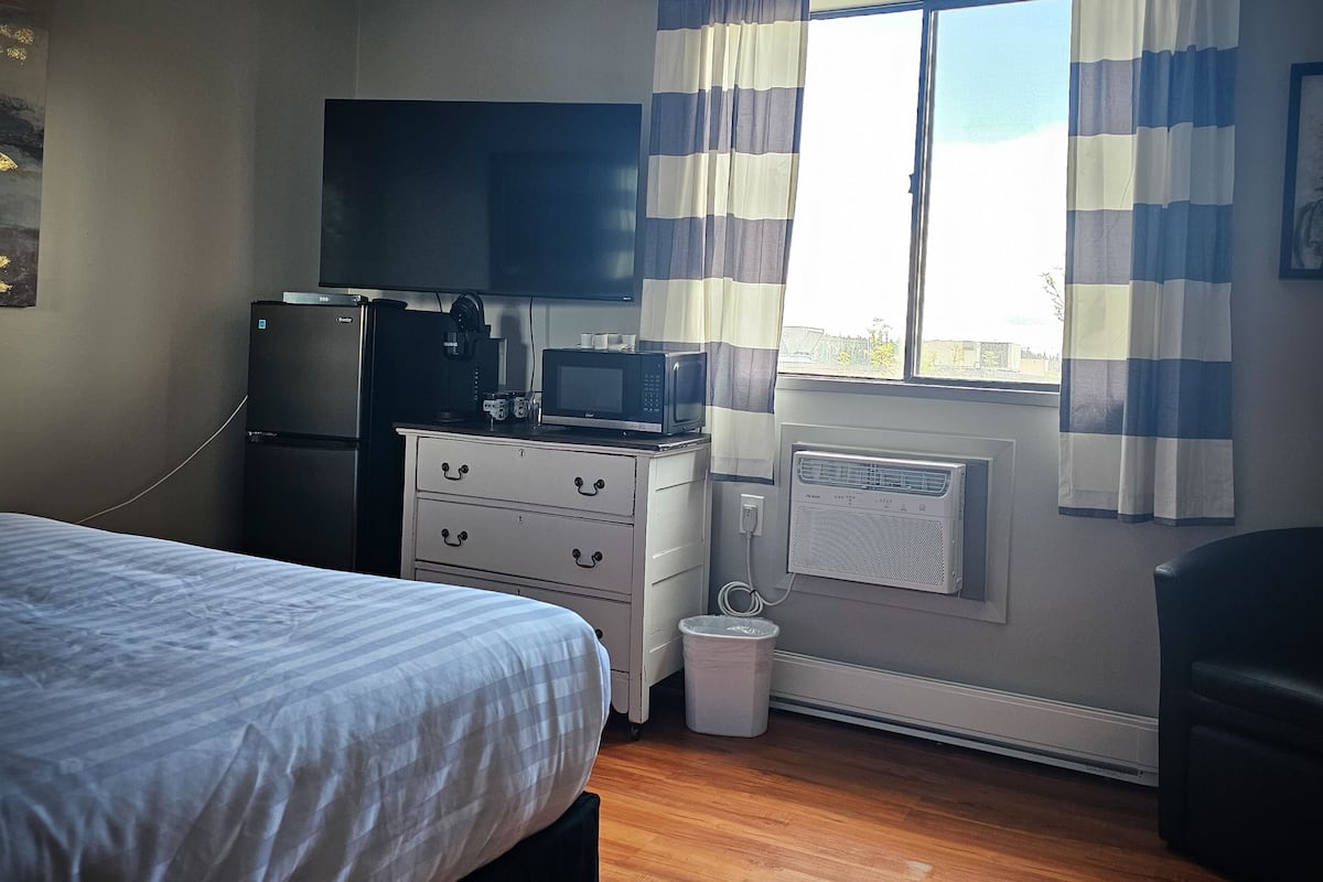A bedroom with a bed, a dresser, a microwave, a television, and a window with curtains.