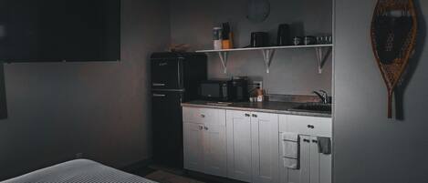 A compact kitchen with a refrigerator, microwave, and coffee maker. There is a shelf above the counter holding kitchenware.