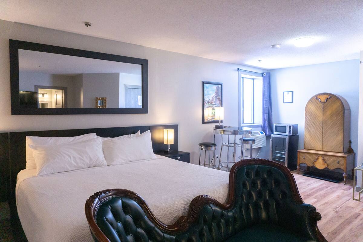 A hotel room with a large bed, a sitting area with a chair, a desk with a chair, a wardrobe, and a large mirror on the wall.