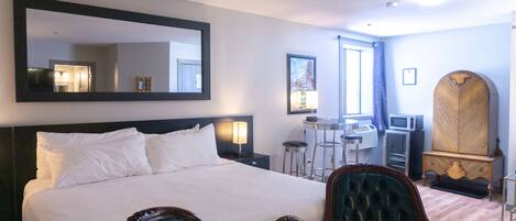 A hotel room with a large bed, a sitting area with a chair, a desk with a chair, a wardrobe, and a large mirror on the wall.