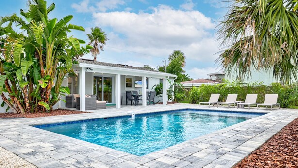 Una piscina con una casa blanca al fondo, rodeada de plantas tropicales.