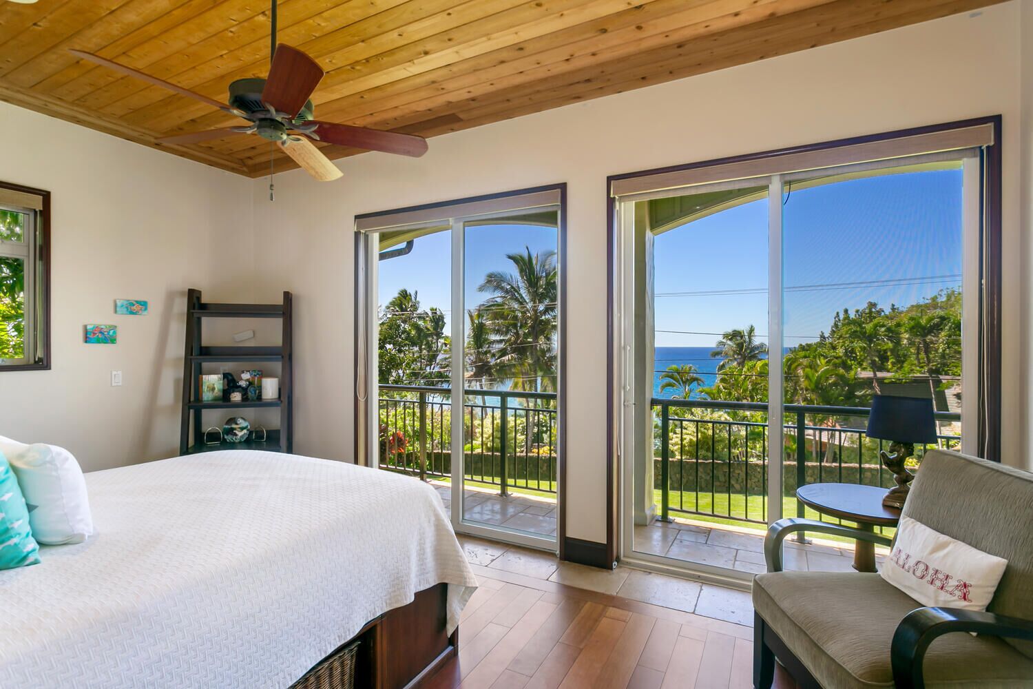 A bedroom with a bed, a chair, a ceiling fan, and a balcony with a view of the ocean and palm trees.