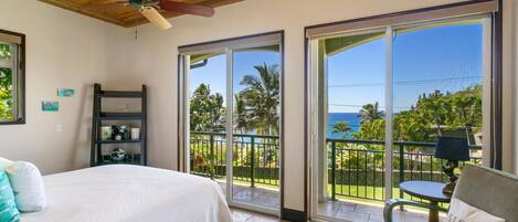 A bedroom with a bed, a chair, a ceiling fan, and a balcony with a view of the ocean and palm trees.