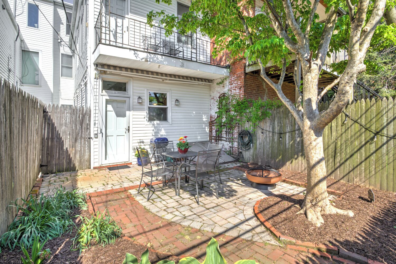 A backyard with a patio, a table, chairs, a fire pit, and a tree.