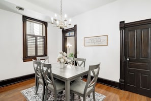 A dining room with a wooden table, grey chairs, a chandelier, a window with blinds, a framed quote on the wall, and a dark wooden door.