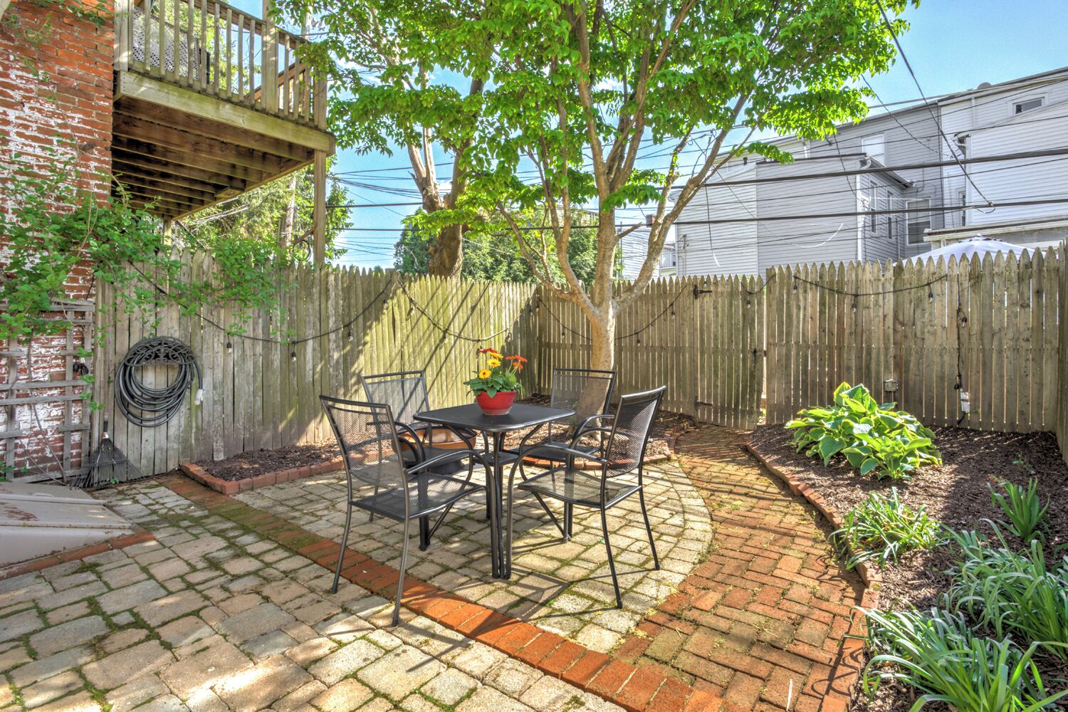 Eine Terrasse mit Tisch und Stühlen, ein gepflasterter Weg, ein Holzzaun und ein Gartenbereich mit Pflanzen.