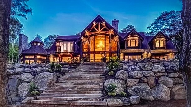 A large, multi-story house with a prominent central tower, stone steps leading to the entrance, and a well-lit facade. - Magnificent 7bdrm 8bath luxury on Lake Joe! Hot Tub! Gym! Boathouse! Sauna! (Seguin)