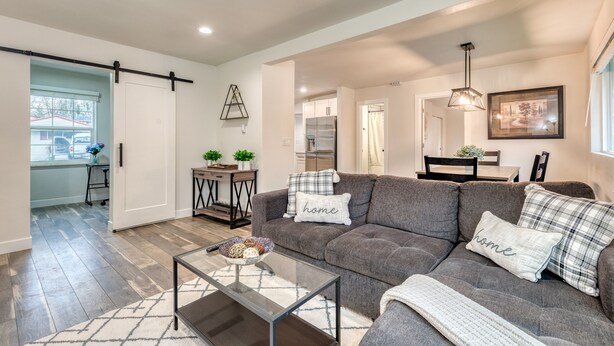 A modern living room with a gray sofa, a glass coffee table, and a dining area in the background. - 1905 Farmhouse w/ Fresh Make Over ☼ 3 BR / 2 BA (Mountlake Terrace)