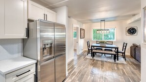 A modern kitchen with stainless steel appliances, a dining area with a wooden table and chairs, and a large window with a view of greenery. - 1905 Farmhouse w/ Fresh Make Over ☼ 3 BR / 2 BA (Mountlake Terrace)