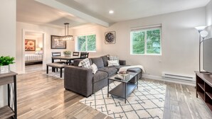 A modern living room with a gray sofa, a coffee table, and a dining area with a table and chairs. There is a large window providing natural light and a view of greenery outside. - 1905 Farmhouse w/ Fresh Make Over ☼ 3 BR / 2 BA (Mountlake Terrace)