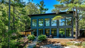 A modern two-story house with large windows, a stone staircase, and a forested backdrop.