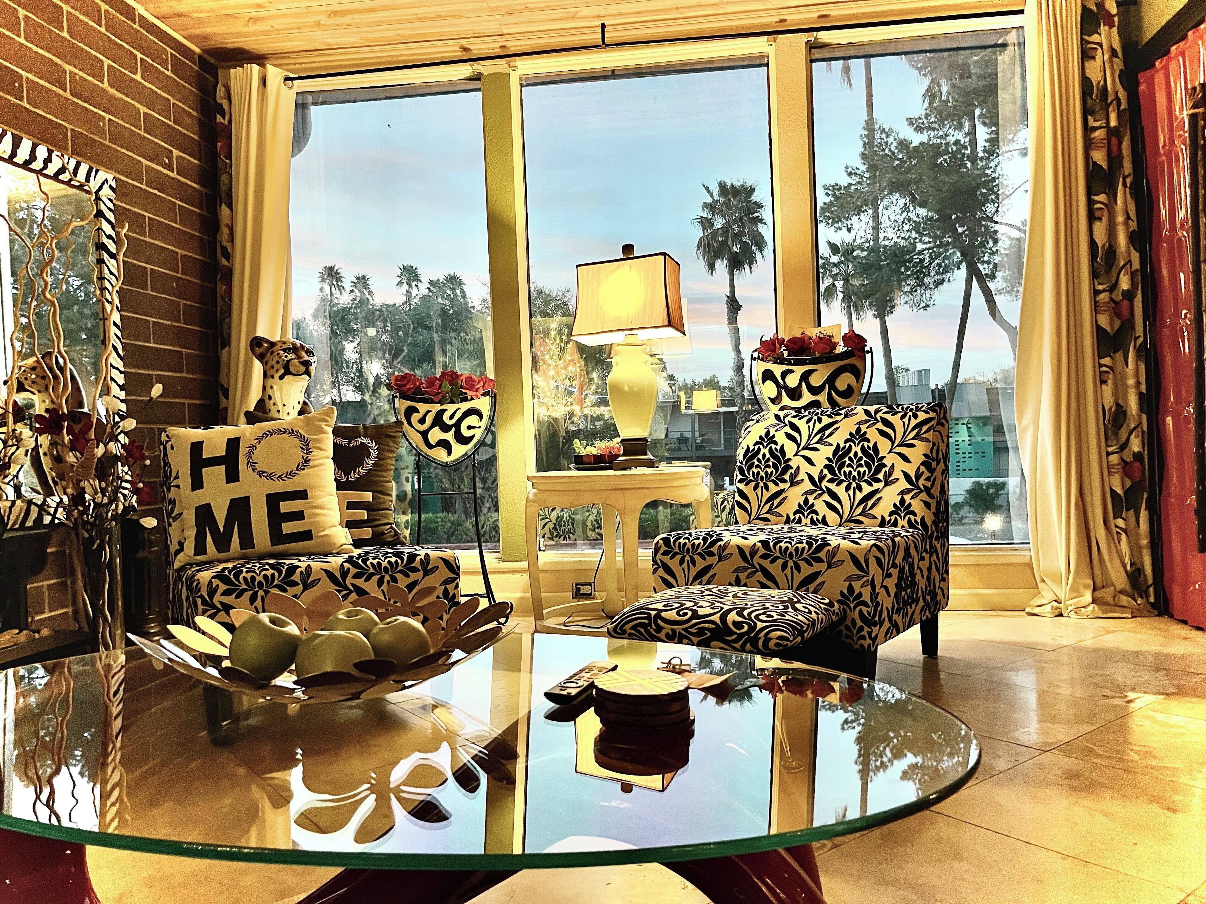 Un salon avec une table basse en verre, un canapé et un fauteuil au motif décoratif. Une fenêtre offre une vue sur des palmiers et le paysage urbain.