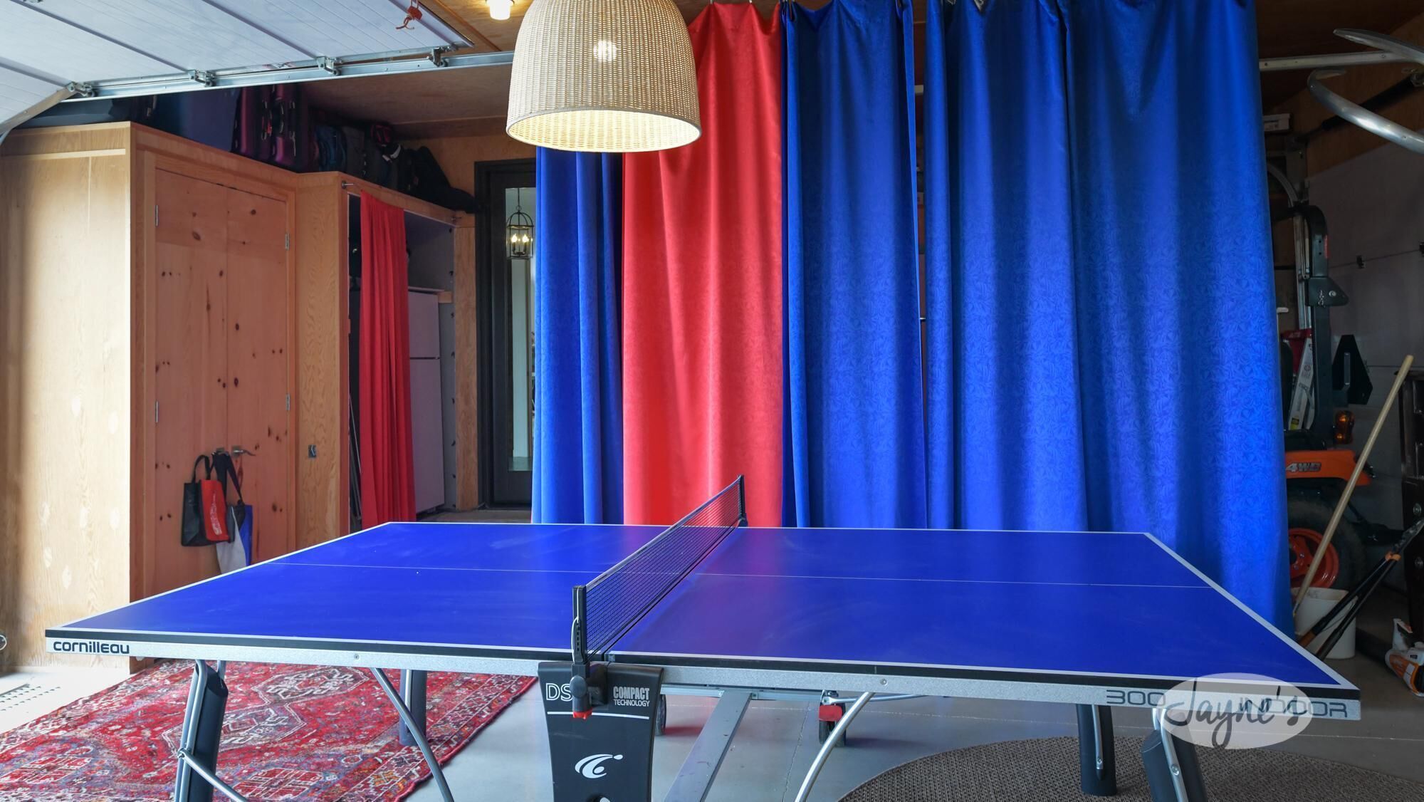 A ping pong table in a room with a wooden cabinet, red and blue curtains, and a hanging light fixture.