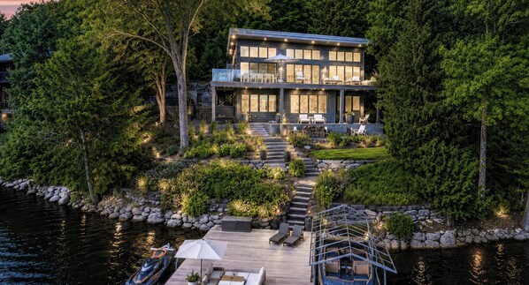 A modern two-story house with a deck, surrounded by lush greenery and a body of water.