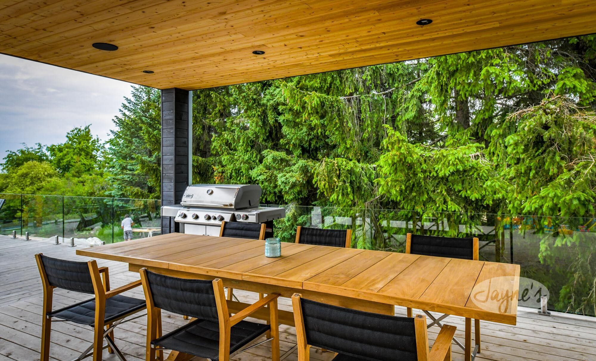 Un área de comedor al aire libre con mesa y sillas de madera, una parrilla y barandilla de cristal con vista a un área boscosa.