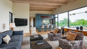 A modern living room with a sofa, coffee table, patterned armchair, and a kitchen area with bar stools. - Lovely 5bdrm 3bath modern home in Creemore! (Creemore)