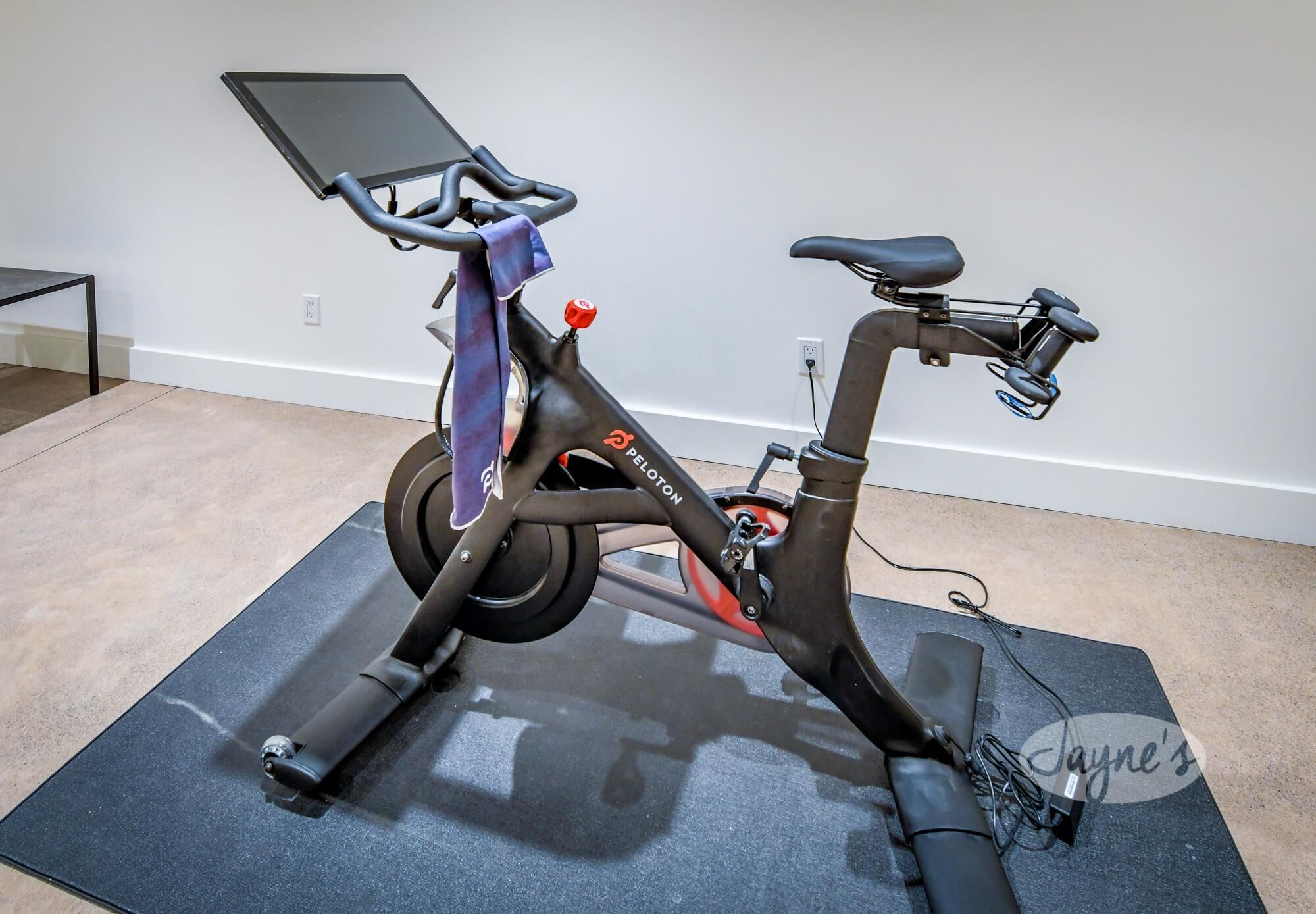 Una bicicleta estática con pantalla digital, ubicada sobre una colchoneta en una habitación con pared lisa y un enchufe.