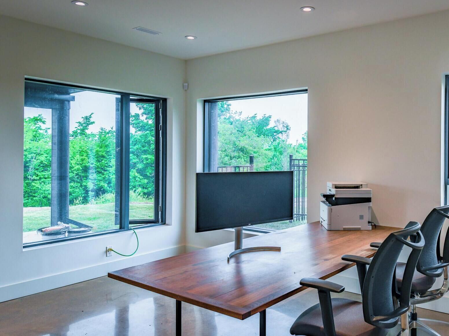 A modern office space with a large desk, a computer monitor, and a printer. There are two large windows providing views of a green landscape.