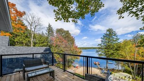 Una terraza de madera con mobiliario de mimbre, una mesa de vidrio y vistas a un lago y árboles.