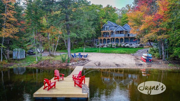 Una propiedad frente al lago con un muelle, sillas Adirondack rojas y una casa grande con porche.
