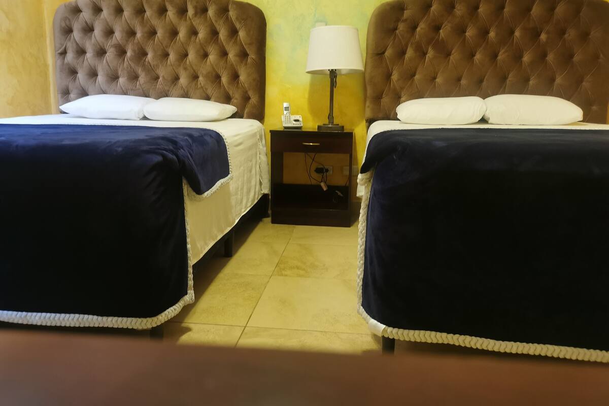 A hotel room with two beds, a nightstand with a lamp, and a yellow wall.