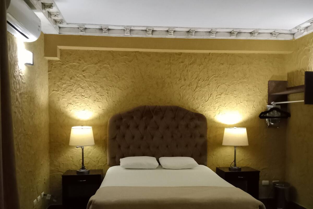 A hotel room with a large bed, two bedside lamps, a wall-mounted heater, and a trash bin.