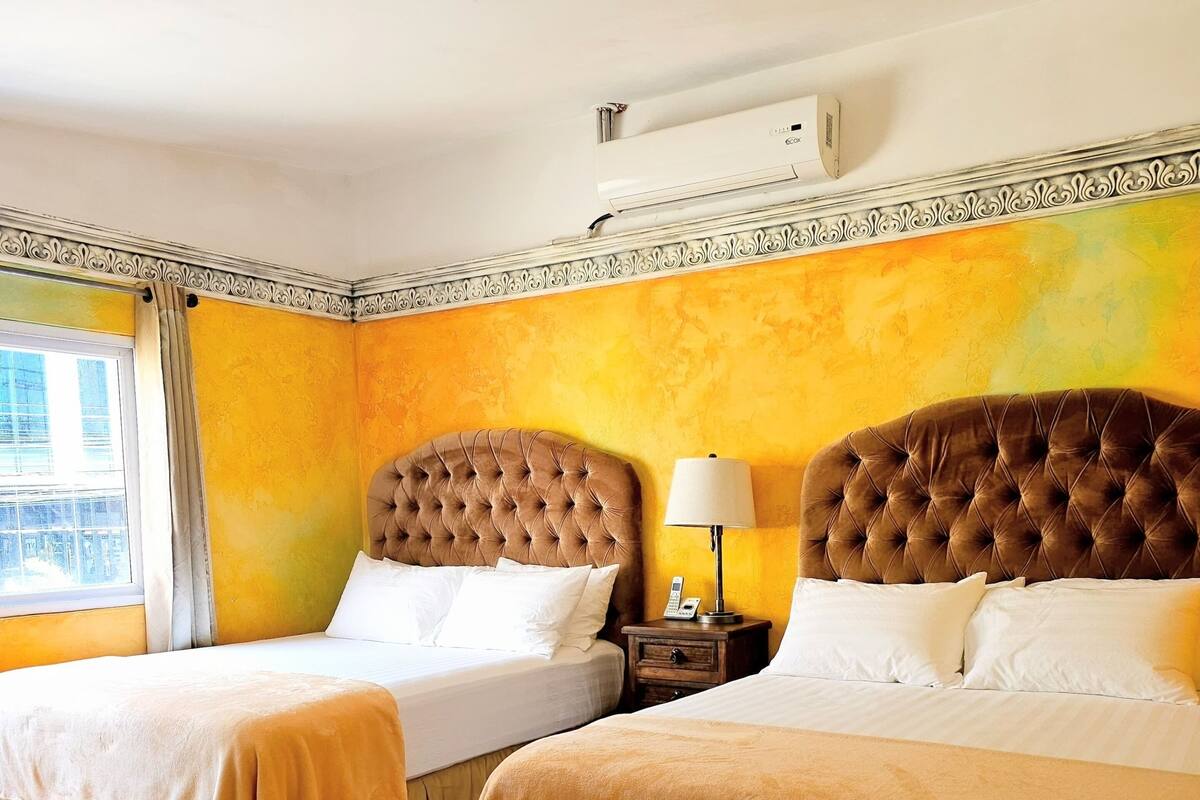 A hotel room with two beds, a window with curtains, a bedside table with a lamp, and an air conditioning unit.
